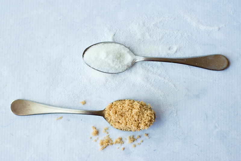 Two spoons laying flat, one filled with brown sugar, and the other filled with white granulated sugar, representing two processed forms of sugar to avoid.